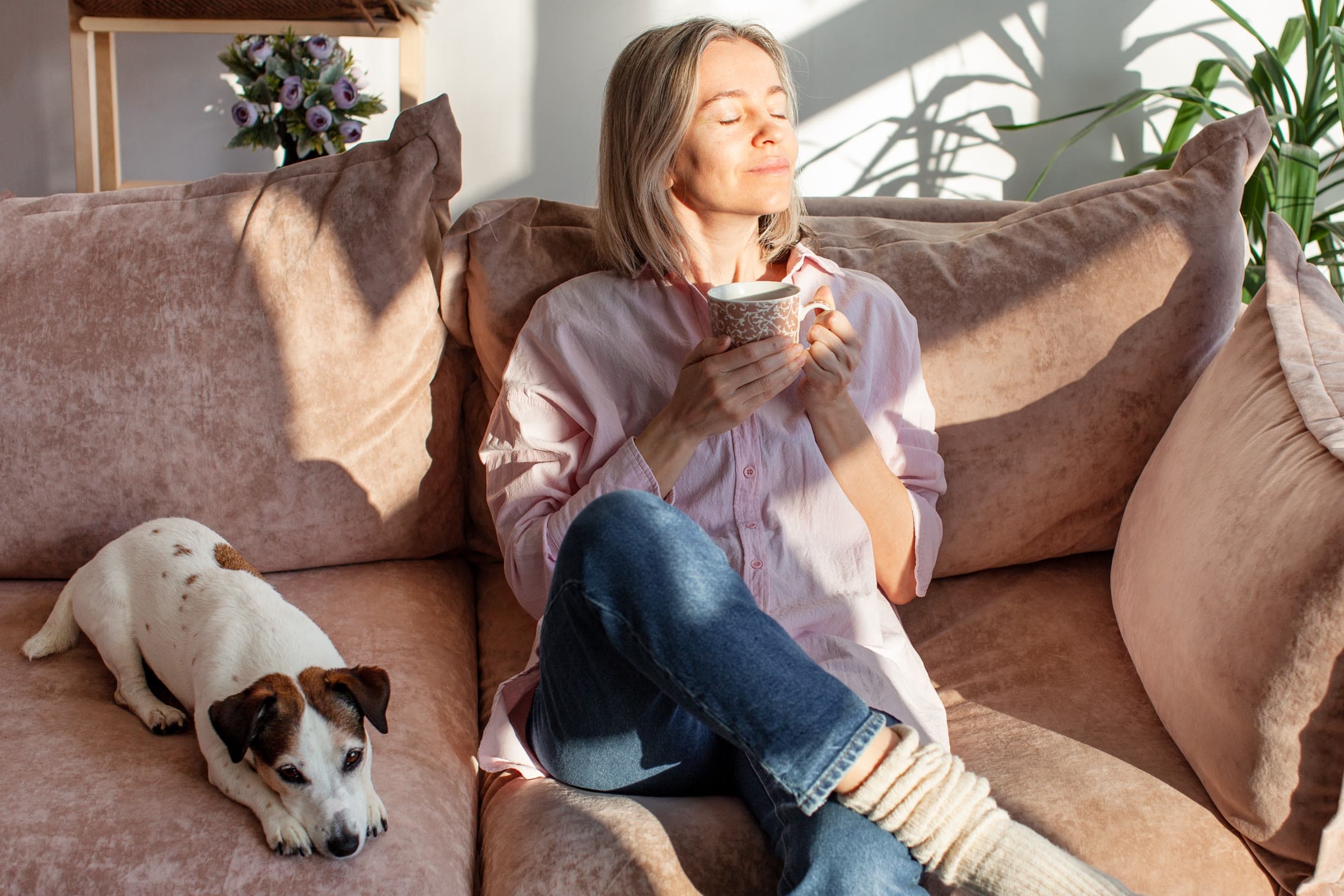 A woman sitting on a sofa in morning sun, eyes closed, holding a mug, with a small dog beside her.