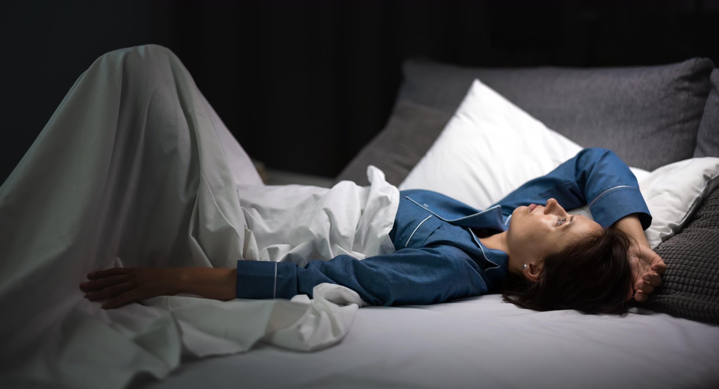 A woman lying awake in bed at night, one hand on her forehead, her knee drawn up under the duvet.